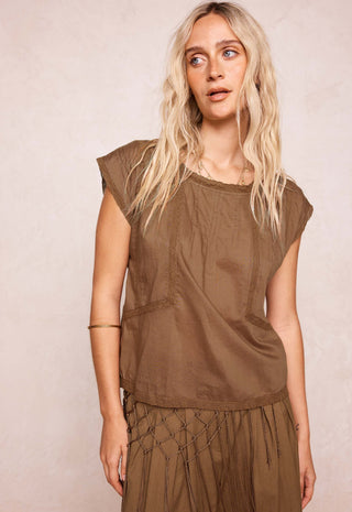 a-woman-with-long-blonde-hair-wearing-a-brownish-olive-brown-sleeveless-top-and-matching-bottoms-standing-against-a-textured-light-beige-wall