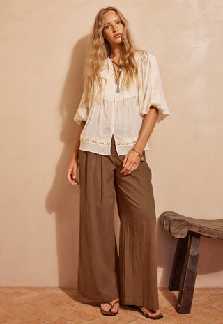 a-woman-stands-in-a-warm-toned-room-wearing-a-loose-white-blouse-with-lace-trim-and-wide-leg-brown-pants-paired-with-sandals-a-rustic-stool-is-visible-against-a-textured-beige-wall