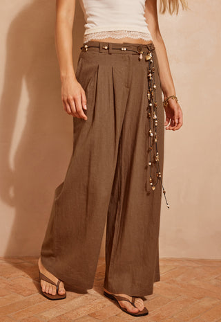 a-model-wearing-high-waisted-wide-leg-brown-pants-with-a-decorative-beaded-belt-and-a-white-lace-trimmed-top-standing-against-a-warm-beige-wall