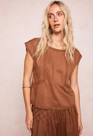 a-woman-with-long-blonde-hair-wearing-a-brown-sleeveless-top-and-matching-brown-skirt-standing-against-a-textured-light-beige-background