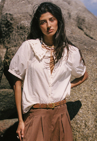 a-woman-wearing-a-white-linen-shirt-with-lace-trim-brown-pants-and-a-studded-belt-accessorized-with-a-chunky-gold-necklace