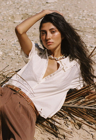 a-woman-reclining-outdoors-on-a-sandy-ground-wearing-a-white-linen-shirt-with-lace-trim-brown-pants-and-a-studded-belt-accessorized-with-a-chunky-gold-necklace