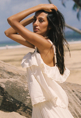 a-woman-on-a-sandy-beach-wearing-a-white-ruffled-sleeveless-dress-with-her-arms-raised-to-her-head-with-the-ocean-and-sky-blurred-in-the-background