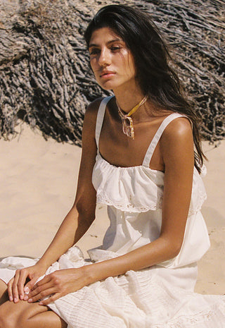a-woman-sitting-on-a-sandy-beach-wearing-a-white-ruffled-spaghetti-strap-dress-and-a-gold-pendant-necklace