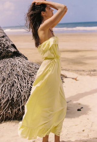 a-woman-stands-on-a-sandy-beach-wearing-a-strapless-pale-yellow-maxi-dress-with-a-gathered-waist-and-ruffled-hem-a-large-thatched-palm-tree-and-ocean-are-visible-in-the-background