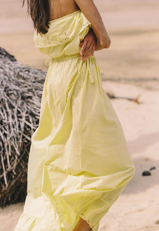 a-woman-wearing-a-flowing-pale-yellow-maxi-dress-standing-on-a-sandy-beach-next-to-a-thatched-structure