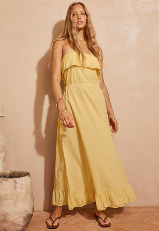 a-woman-modeling-a-strapless-yellow-maxi-dress-with-ruffle-details-standing-against-a-textured-beige-wall-in-a-warm-rustic-setting