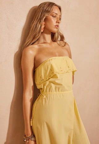 a-woman-modeling-a-strapless-yellow-maxi-dress-with-ruffle-details-standing-against-a-textured-beige-wall-in-a-warm-rustic-setting