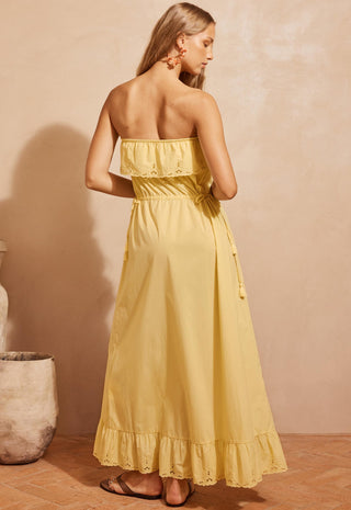 a-woman-modeling-a-strapless-yellow-maxi-dress-with-ruffle-details-standing-against-a-textured-beige-wall-in-a-warm-rustic-setting