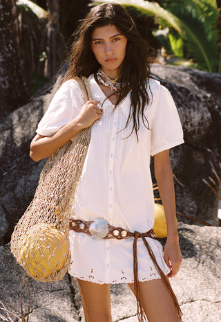 a-woman-stands-outdoors-on-rocky-ground-wearing-a-white-short-sleeve-button-front-shirt-dress-with-eyelet-trim-a-brown-woven-belt-with-a-large-silver-buckle-layered-necklaces-and-a-large-beige-mesh-tote-bag-containing-yellow-coconuts