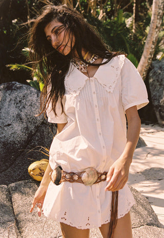 a-woman-wearing-a-white-short-sleeve-bohemian-style-mini-dress-with-eyelet-lace-details-a-button-down-front-and-a-wide-brown-woven-belt-with-large-shell-like-accents-standing-outdoors-on-a-rocky-tropical-path