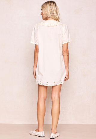 a-woman-shown-from-the-back-wearing-a-short-sleeve-cream-colored-tunic-dress-with-lace-trim-at-the-collar-and-hem-paired-with-white-lace-up-flats-set-against-a-textured-beige-studio-backdrop