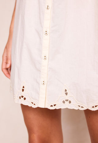close-up-of-a-white-eyelet-button-front-dress-or-tunic-with-a-scalloped-embroidered-hem-shown-on-a-model-against-a-plain-light-beige-background