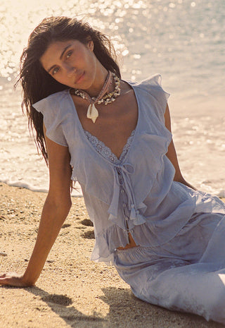 a-woman-sits-on-a-sandy-beach-wearing-a-light-blue-sheer-ruffled-two-piece-outfit-with-lace-trim-and-a-matching-necklace