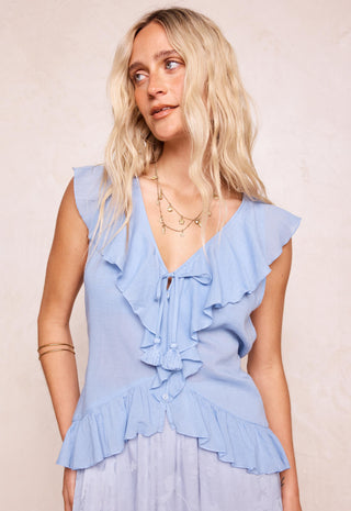 a-blonde-woman-wearing-a-light-blue-sleeveless-ruffled-blouse-with-a-tie-front-detail-and-a-matching-light-blue-skirt-posed-against-a-neutral-beige-background