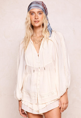 a-woman-wearing-a-cream-colored-long-sleeved-button-up-blouse-with-pleated-details-and-a-matching-tiered-skirt-styled-with-a-blue-patterned-headscarf-against-a-textured-beige-background