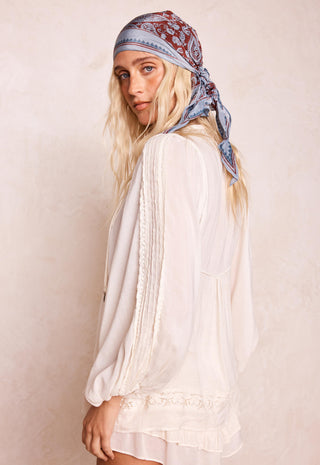 a-woman-wearing-a-white-long-sleeve-lace-trim-blouse-and-a-blue-paisley-bandana-headscarf-photographed-against-a-textured-beige-wall