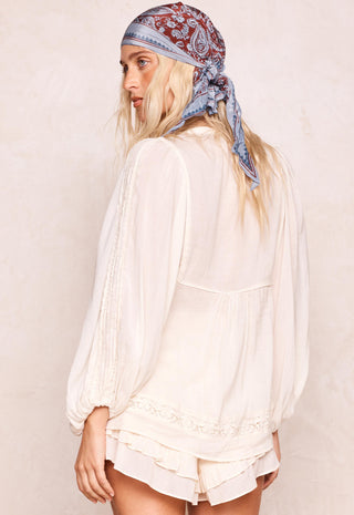 a-woman-shown-from-the-back-wearing-a-cream-colored-long-sleeve-blouse-with-lace-trim-and-matching-ruffled-shorts-paired-with-a-blue-and-red-paisley-bandana-headscarf-against-a-textured-beige-wall