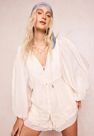 a-woman-modeling-a-cream-colored-bohemian-style-two-piece-set-consisting-of-a-long-sleeve-button-up-top-and-matching-shorts-styled-with-a-blue-patterned-headband-against-a-textured-beige-wall