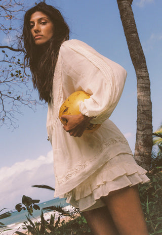 a-woman-wearing-a-loose-white-long-sleeve-mini-dress-with-lace-trim-stands-outdoors-near-a-palm-tree-holding-a-yellow-fruit-against-a-blue-sky-background