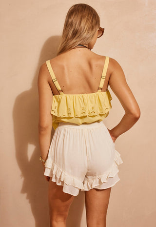 a-woman-shown-from-the-back-wearing-a-pale-yellow-ruffled-eyelet-camisole-top-with-suspenders-and-white-ruffled-shorts-standing-against-a-warm-beige-wall