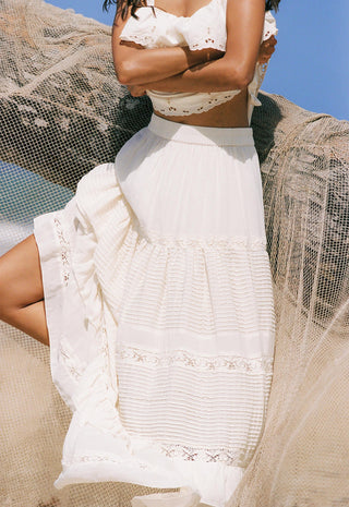 a-woman-wearing-a-white-embroidered-maxi-skirt-and-matching-white-crop-top-posed-outdoors-against-a-blue-sky-with-sheer-beige-fabric-draped-around-her