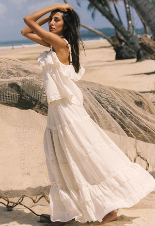 A woman wearing a long white tiered boho maxi dress stands on a sandy beach next to a large driftwood log