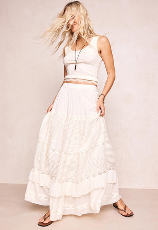 a-woman-modeling-a-white-sleeveless-crop-top-and-a-long-white-tiered-skirt-with-embroidered-details-paired-with-brown-sandals-against-a-neutral-beige-studio-backdrop