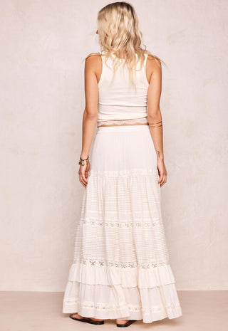 a-woman-shown-from-the-back-wearing-a-sleeveless-cream-top-and-a-long-cream-tiered-skirt-with-lace-trim-standing-against-a-light-beige-textured-wall