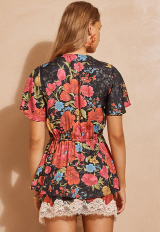 a-woman-shown-from-the-back-wearing-a-short-sleeve-floral-romper-with-a-black-base-and-vibrant-pink-red-blue-and-yellow-flowers-featuring-a-lace-hem