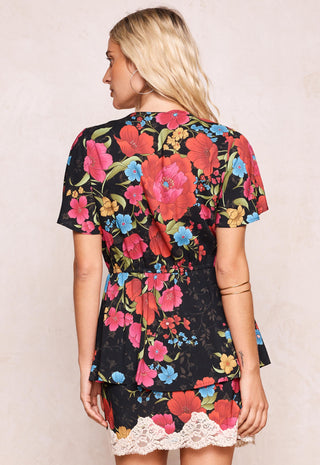 a-woman-shown-from-the-back-wearing-a-short-sleeve-black-dress-with-a-vibrant-red-pink-and-blue-floral-print-and-a-white-lace-hem-set-against-a-plain-beige-background