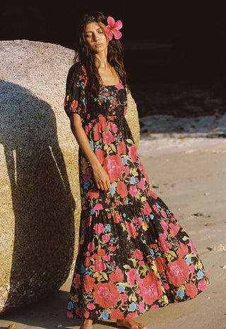 a-woman-stands-barefoot-on-a-sandy-beach-beside-a-large-rock-wearing-a-black-maxi-dress-with-a-vibrant-pink-red-and-blue-floral-print-she-has-a-pink-flower-tucked-into-her-dark-hair