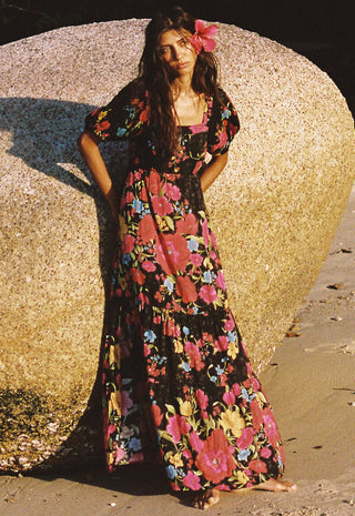 a-woman-stands-barefoot-outdoors-against-a-large-rock-wearing-a-long-black-maxi-dress-with-a-vibrant-pink-red-and-blue-floral-print-and-a-pink-flower-in-her-hair