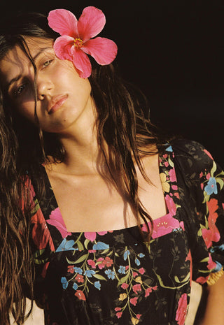 a-woman-with-long-dark-hair-and-a-pink-flower-on-her-head-wearing-a-black-floral-top-with-pink-blue-and-yellow-flowers-against-a-dark-background