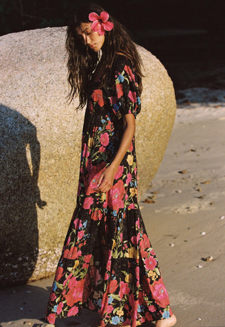 a-woman-stands-on-a-sandy-beach-beside-a-large-rock-wearing-a-long-black-maxi-dress-with-a-vibrant-pink-red-and-blue-floral-print-she-has-a-pink-flower-in-her-hair