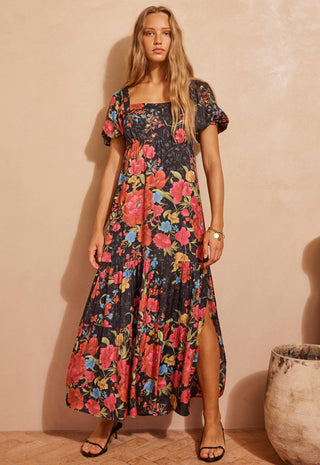 a-woman-modeling-a-black-maxi-dress-with-a-vibrant-red-pink-and-blue-floral-print-standing-against-a-textured-beige-wall