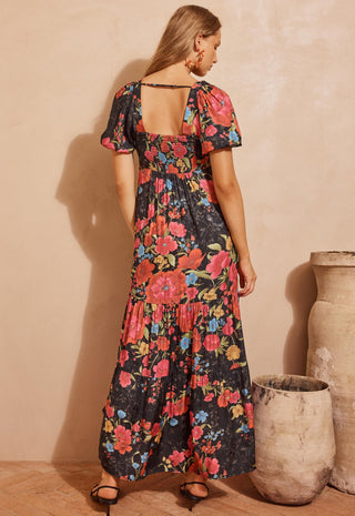 a-woman-wearing-a-black-maxi-dress-with-a-vibrant-pink-red-and-blue-floral-print-shown-from-the-back-against-a-warm-beige-textured-wall-with-rustic-clay-pots