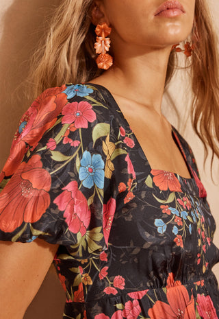 a-woman-modeling-a-black-maxi-dress-with-a-vibrant-red-pink-and-blue-floral-print-standing-against-a-textured-beige-wall