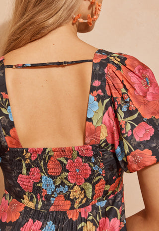 a-back-shot-of-a-woman-modeling-a-black-maxi-dress-with-a-vibrant-red-pink-and-blue-floral-print-standing-against-a-textured-beige-wall