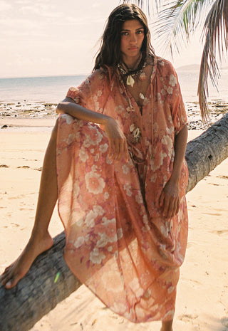a-woman-on-a-sandy-beach-sits-on-a-fallen-palm-tree-wearing-a-flowing-peach-floral-kaftan-dress-the-background-shows-the-ocean-sand-and-palm-leaves