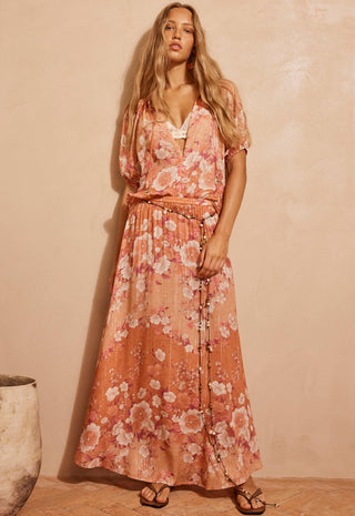 a-woman-modeling-a-peach-and-pink-floral-maxi-dress-with-a-tie-waist-and-short-flutter-sleeves-standing-against-a-textured-beige-wall