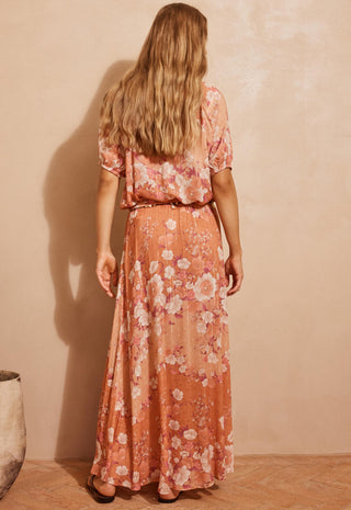 a-woman-viewed-from-behind-wearing-a-peach-orange-maxi-dress-with-a-pink-and-white-floral-print-standing-against-a-textured-beige-wall