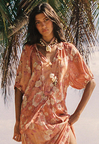 a-woman-wearing-a-loose-peach-floral-cover-up-dress-with-shell-like-tie-details-standing-outdoors-near-palm-leaves-against-a-bright-beach-like-background