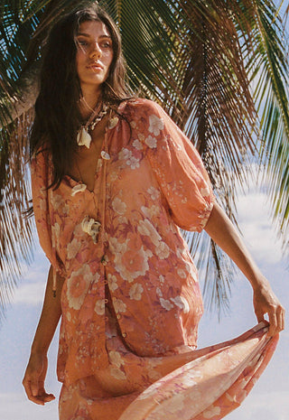 a-woman-wearing-a-loose-short-sleeve-peach-floral-cover-up-dress-with-a-tie-neckline-standing-outdoors-under-palm-leaves-against-a-bright-sky