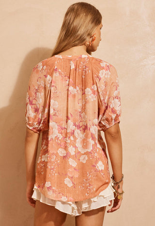 a-woman-shown-from-the-back-wearing-a-peach-colored-short-sleeve-blouse-with-a-pink-and-white-floral-print-paired-with-white-ruffled-shorts-set-against-a-textured-beige-wall