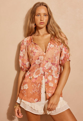a-woman-modeling-a-short-sleeve-button-up-blouse-with-a-peach-pink-and-white-floral-print-paired-with-white-ruffled-shorts-against-a-warm-beige-background