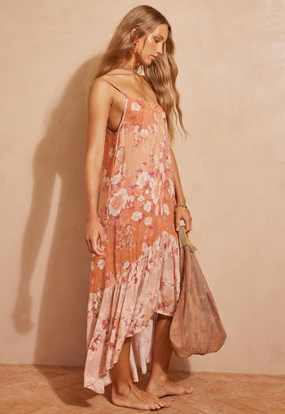 a-woman-stands-barefoot-against-a-warm-beige-wall-wearing-a-peach-and-pink-floral-spaghetti-strap-maxi-dress-and-holding-a-woven-tote-bag