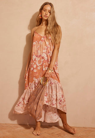 a-woman-modeling-a-sleeveless-high-low-floral-maxi-dress-in-a-peach-and-pink-color-palette-with-a-tie-dye-pattern-standing-against-a-textured-beige-wall