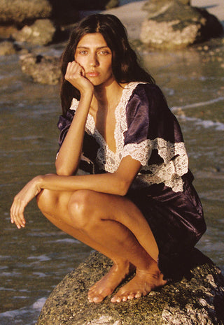 a-woman-crouches-barefoot-on-a-rock-by-the-water-wearing-a-dark-purple-satin-robe-with-white-lace-trim