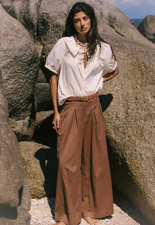 a-woman-stands-outdoors-between-large-rock-formations-wearing-a-loose-white-blouse-with-lace-up-front-details-and-wide-brown-pleated-pants
